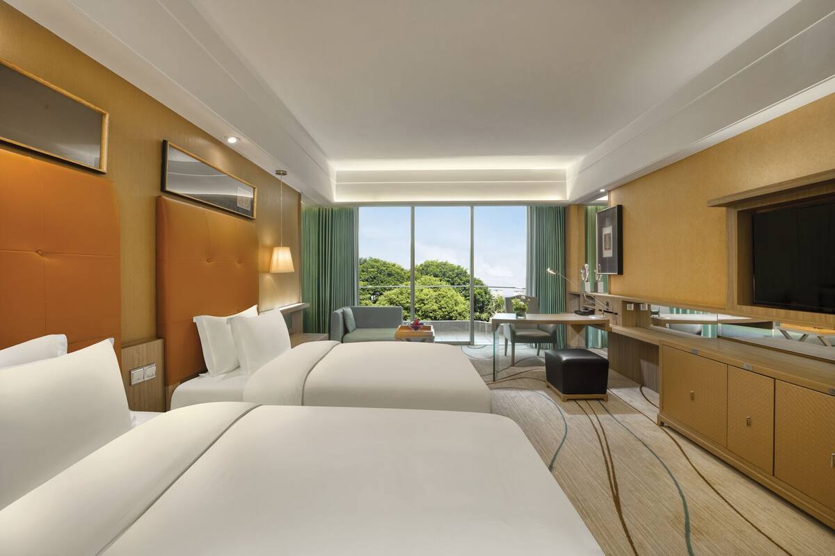 Habitación de hotel con dos camas, un escritorio, una silla, un televisor y un ventanal con vistas a árboles.