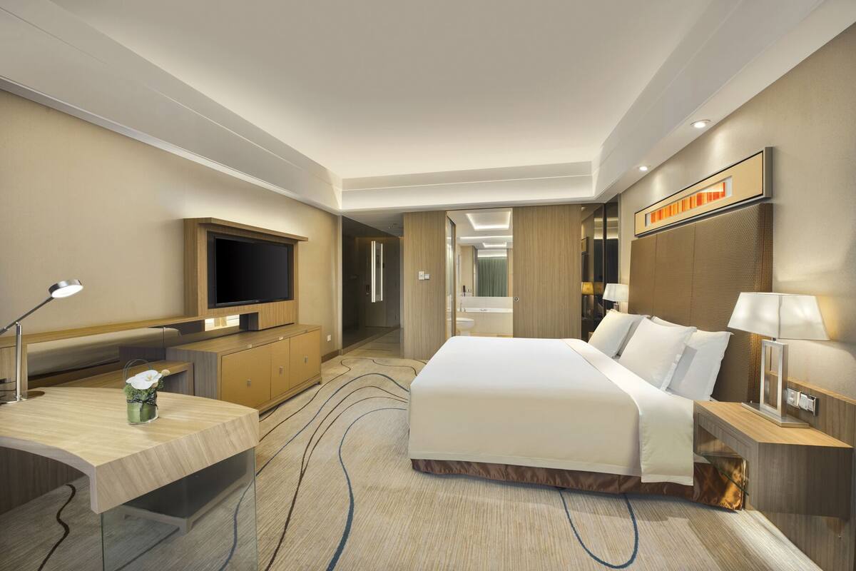 Una habitación de hotel moderna con una cama grande, un escritorio con lámpara, televisión y baño visible a través de una puerta abierta.