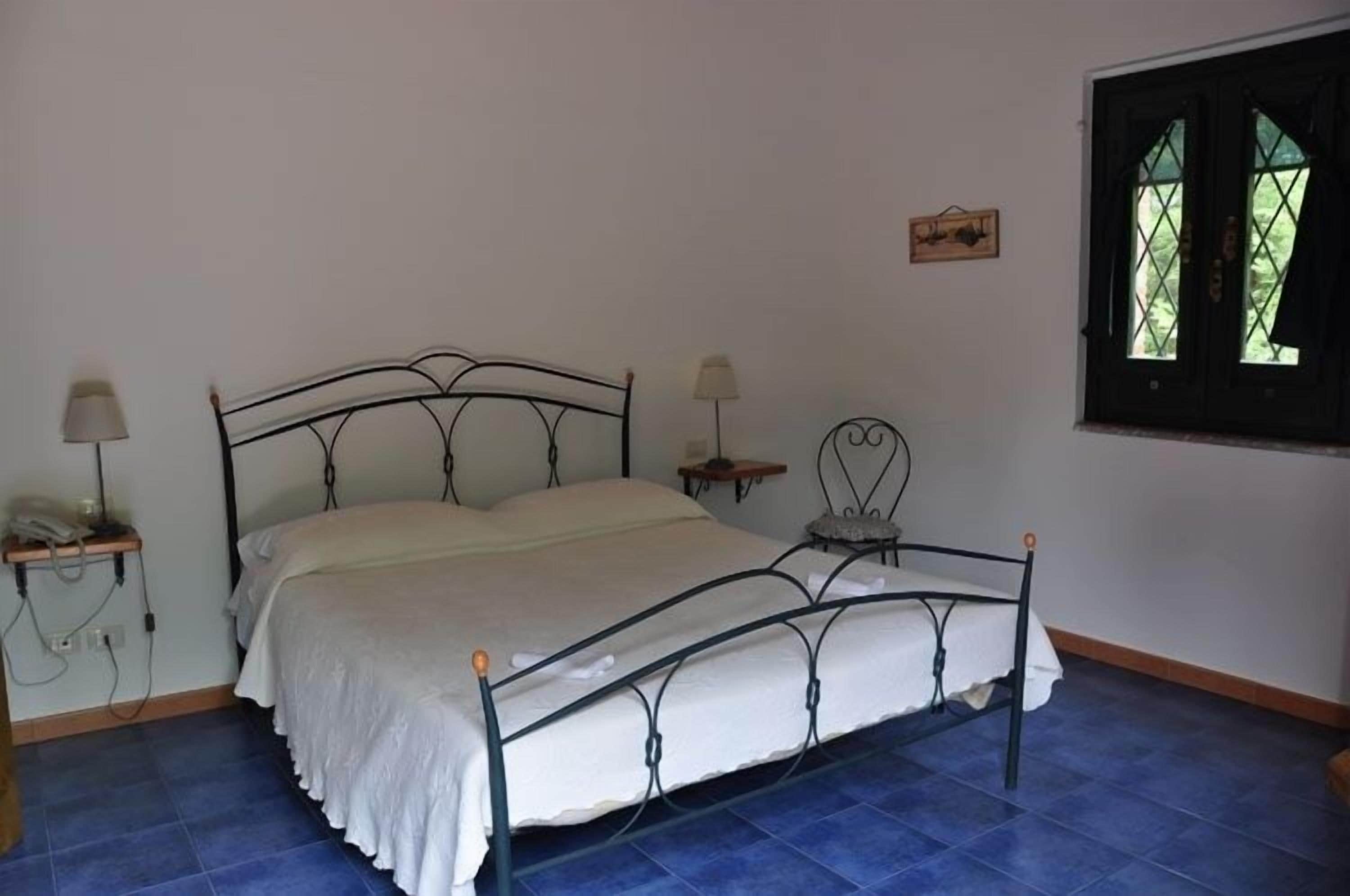 Uma cama de solteiro com estrutura de metal, uma mesinha de cabeceira com abajur e uma janela com vista para o verde.