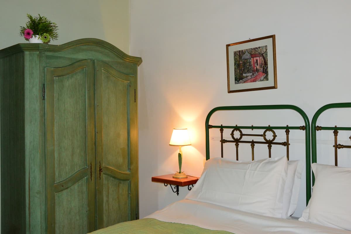 A room with a bed, a nightstand with a lamp, a green metal headboard, and a wooden wardrobe.