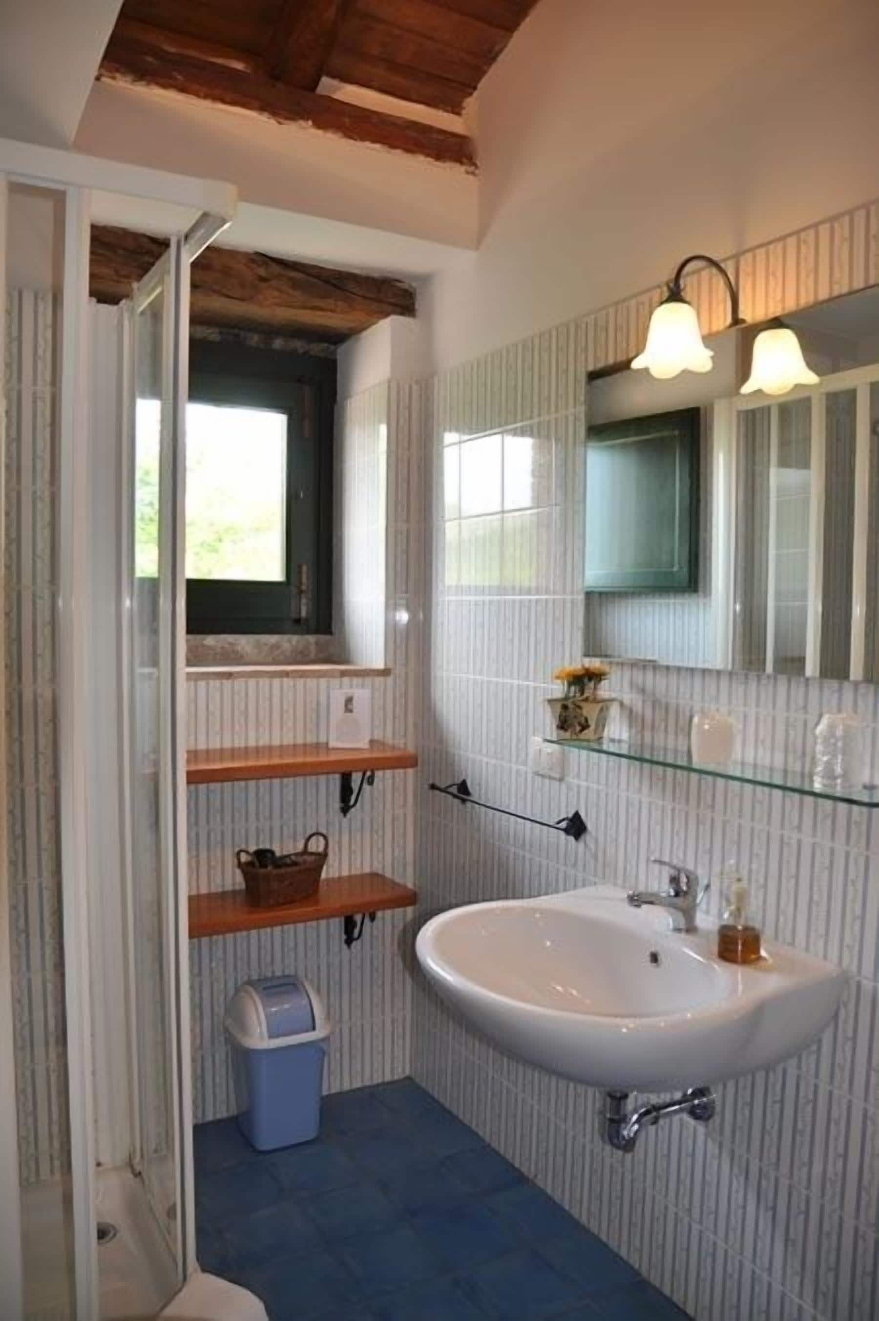 Une salle de bain avec un lavabo blanc, des étagères en bois, une poubelle en verre et une fenêtre donnant sur un beau paysage.