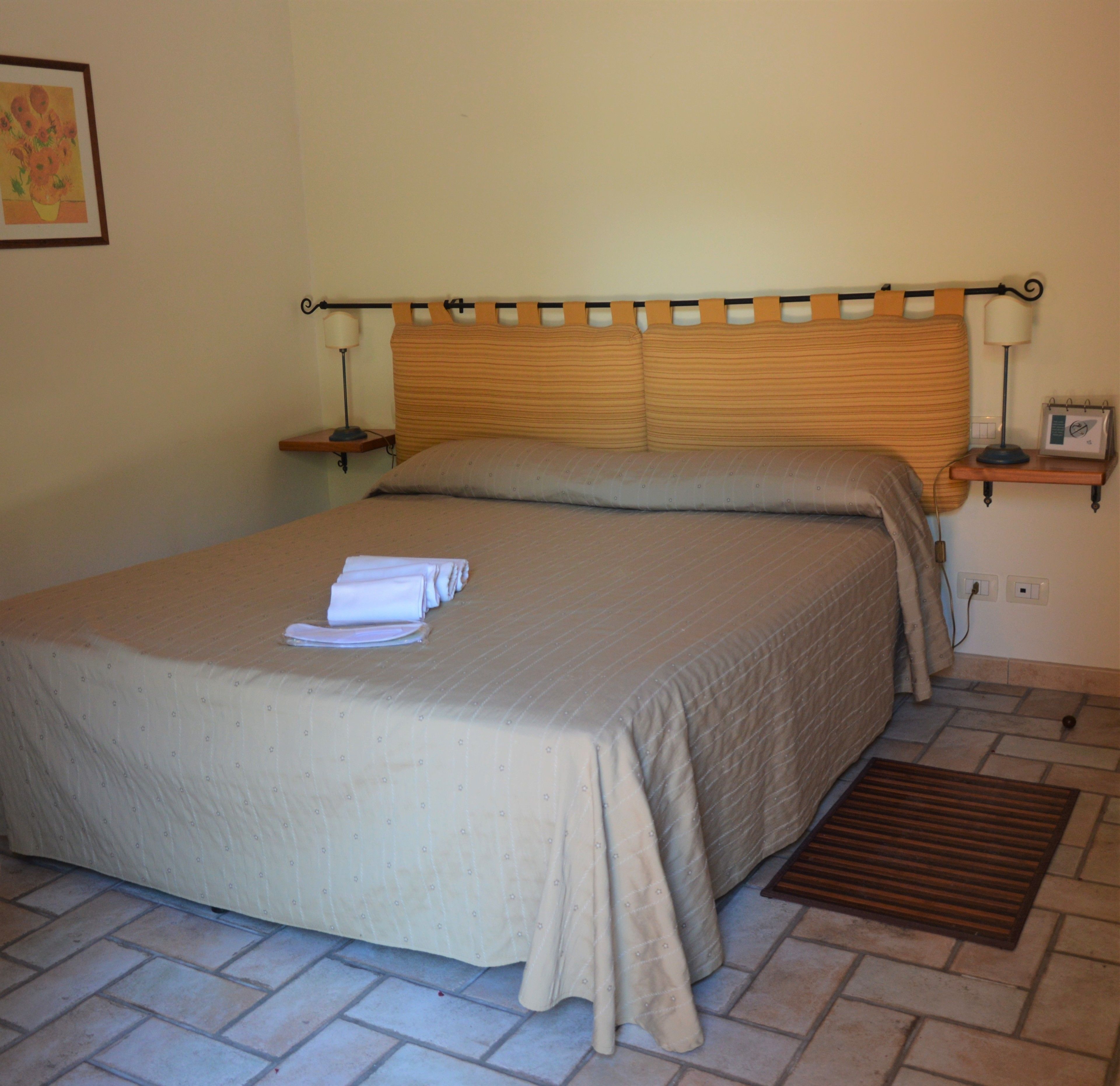 A neatly made bed with a beige bedspread and pillows, a bedside table with a lamp, and a small shelf with a clock and calendar.
