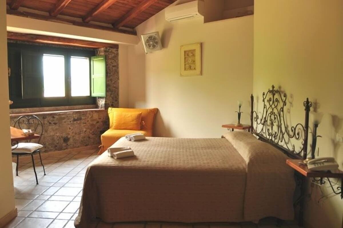 A bedroom with a bed, a chair, a small table, a window with shutters, and a picture on the wall.