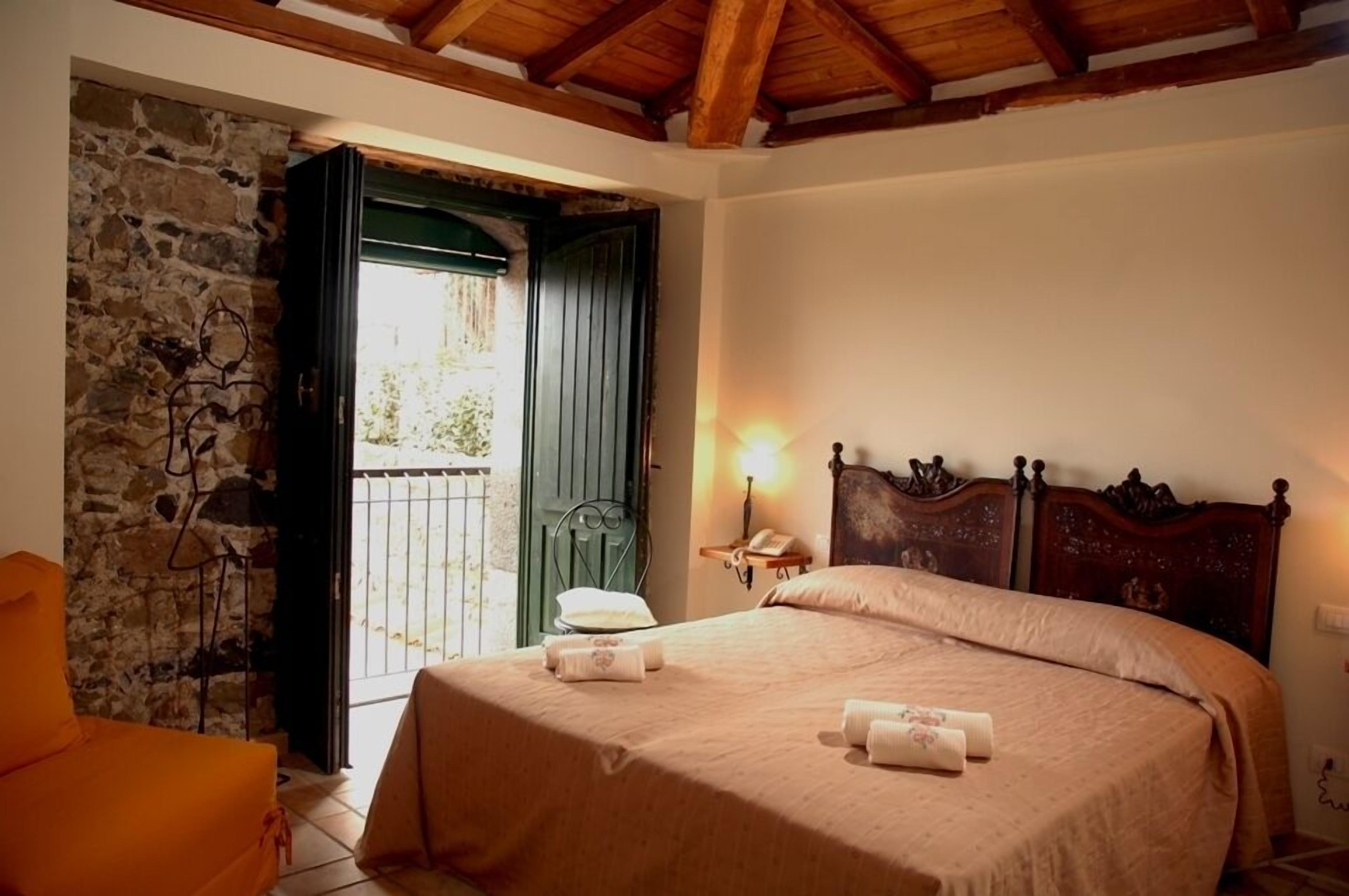 Un dormitorio con pared de piedra, vigas de madera en el techo, una cama con cabecera, una mesita de noche con lámpara y vistas a través de una puerta abierta.