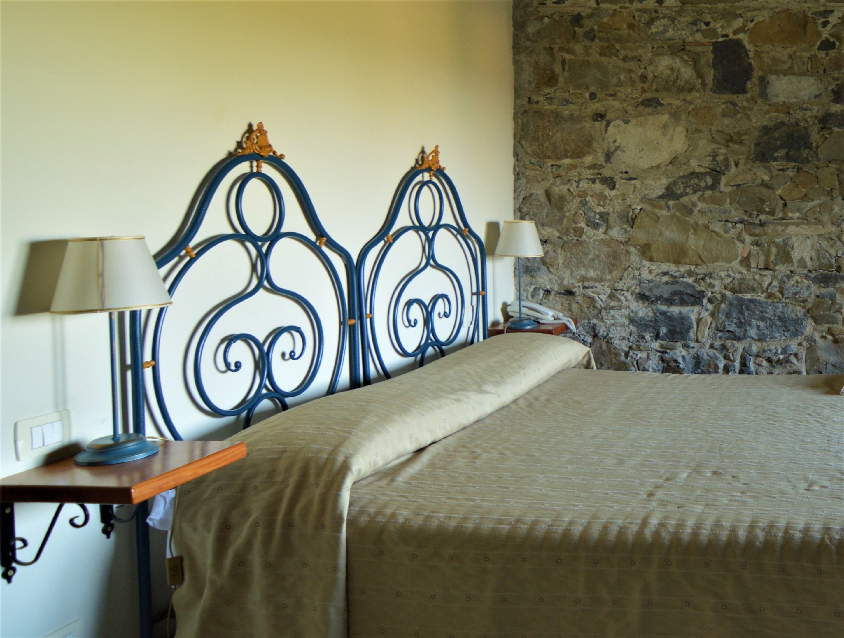 Ein Schlafzimmer mit einer Steinwand, einem Bett mit beiger Bettdecke, zwei Nachttischen mit Lampen und einem dekorativen Metall-Kopfteil.