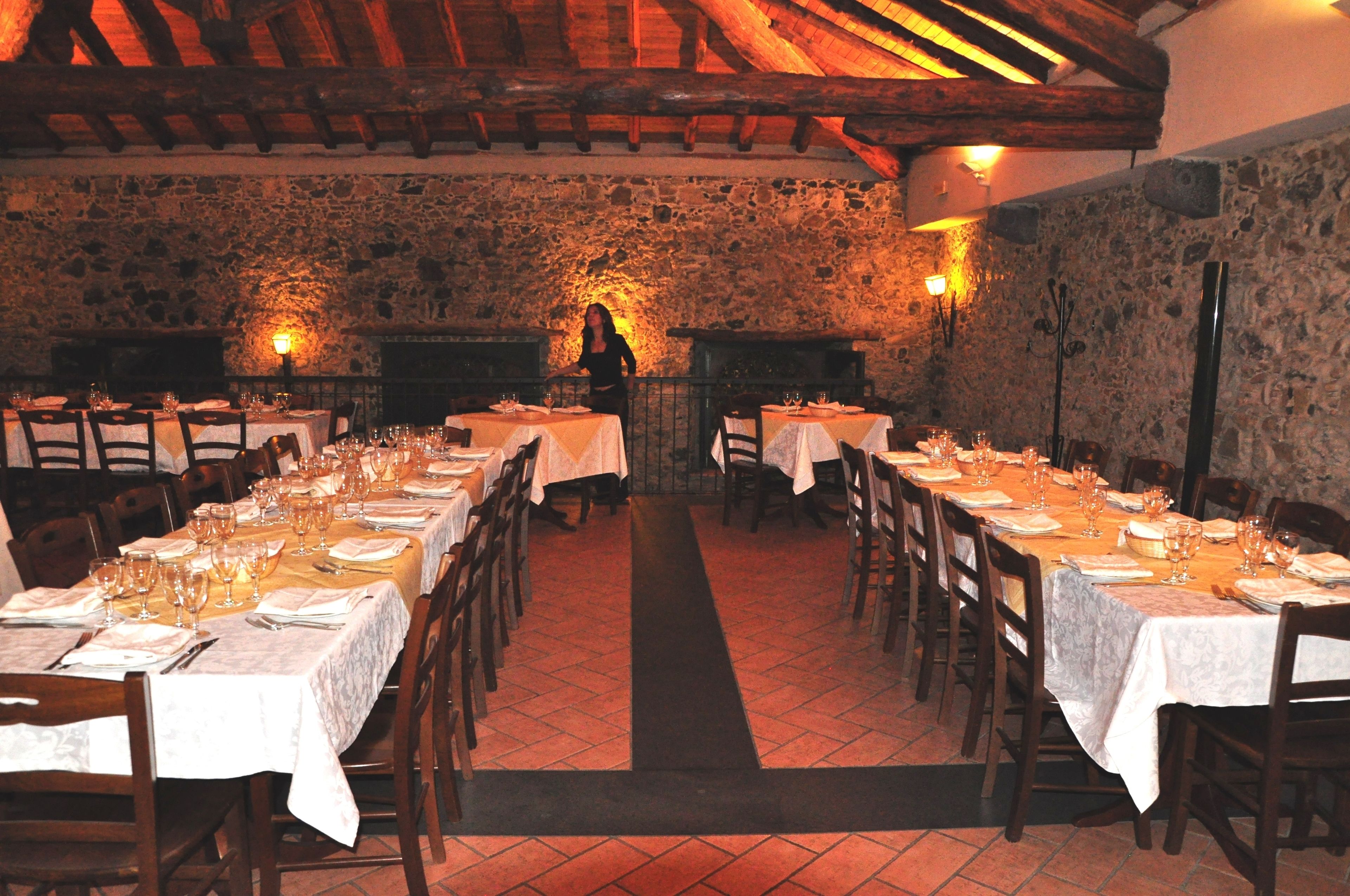 Un restaurante con paredes de piedra y vigas de madera, mesas dispuestas con manteles blancos y copas de vino, y un pasillo central.