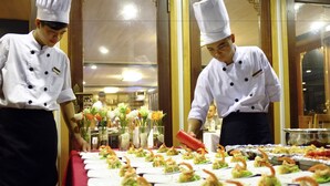 Two chefs preparing food in a professional kitchen with a long table of appetizers. - Halong Glory Legend Cruise (Tuan Chau)