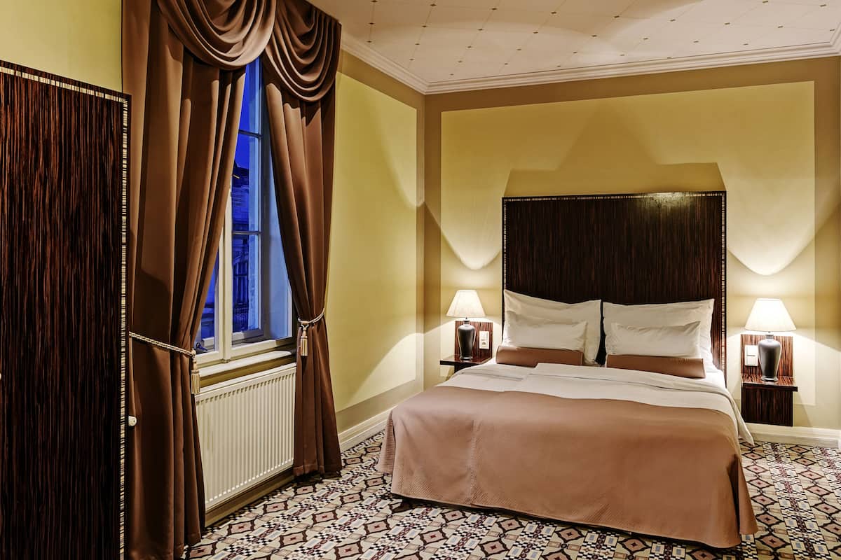 Ein Hotelzimmer mit einem großen Bett, zwei Nachttischen mit Lampen, einem Fenster mit Vorhängen und einem gemusterten Boden.