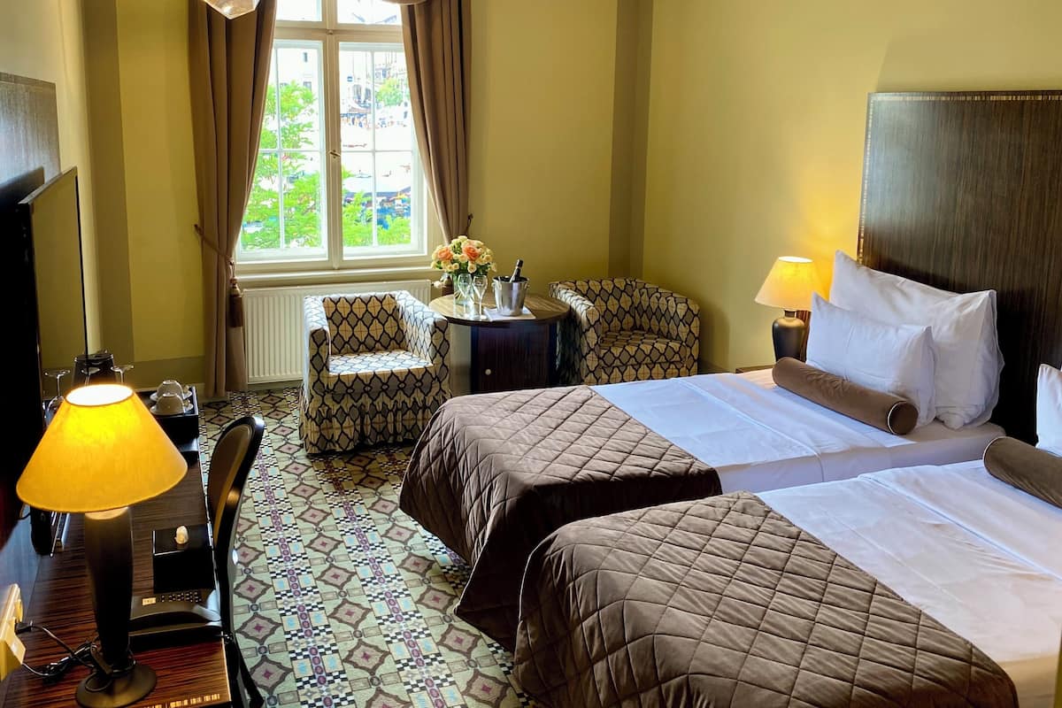 Ein Hotelzimmer mit Bett, zwei Stühlen, einem Schreibtisch mit Lampe, einem Fenster mit Vorhängen und Blick ins Freie.