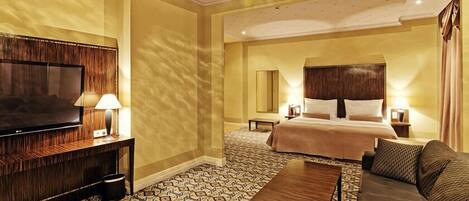 A hotel room with a large flat-screen TV, a sofa, a bed with a wooden headboard, and patterned floor tiles.