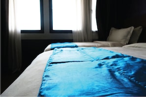 Ein ordentlich bezogenes Bett mit einer blauen Decke, ein Fenster mit Vorhängen und eine dunkle Wand.