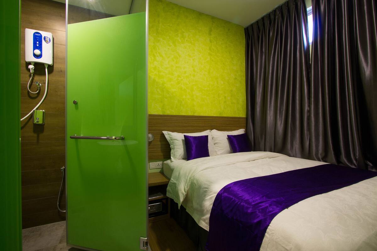 Een hotelkamer met een groene garderobe, een bed met wit en paars beddengoed, een houten hoofdeinde en een douche met bedieningspaneel.