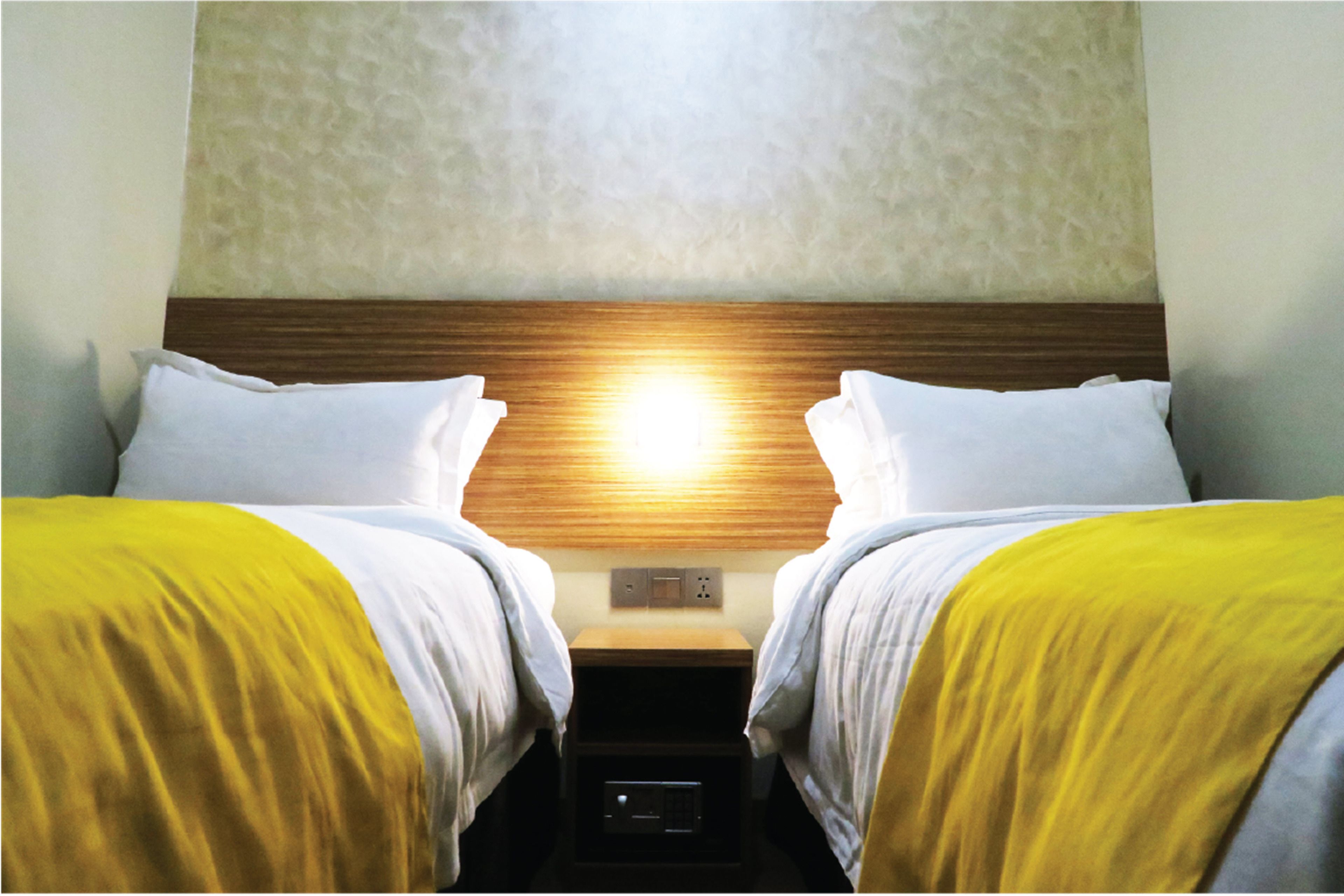 A hotel room with two beds, each with white bedding and a yellow throw blanket. There is a wooden headboard and a nightstand with a lamp between the beds.