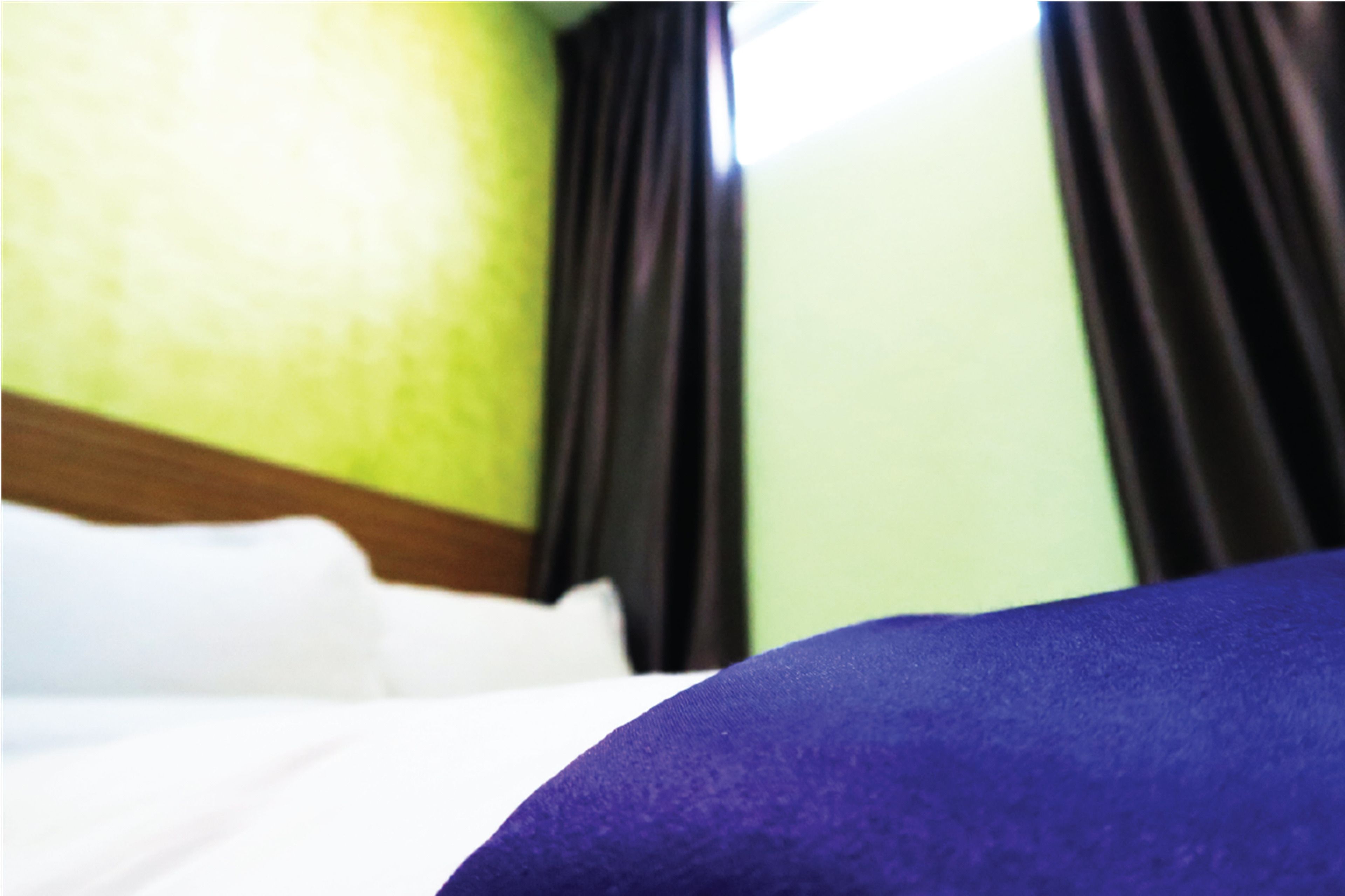 Un lit avec du linge de lit blanc et un oreiller violet, un mur vert et des rideaux foncés.