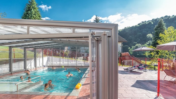 An indoor swimming pool with clear glass walls, surrounded by a red fence and a covered patio area. - Hôtel et Restaurant du Col Osquich (Musculdy)
