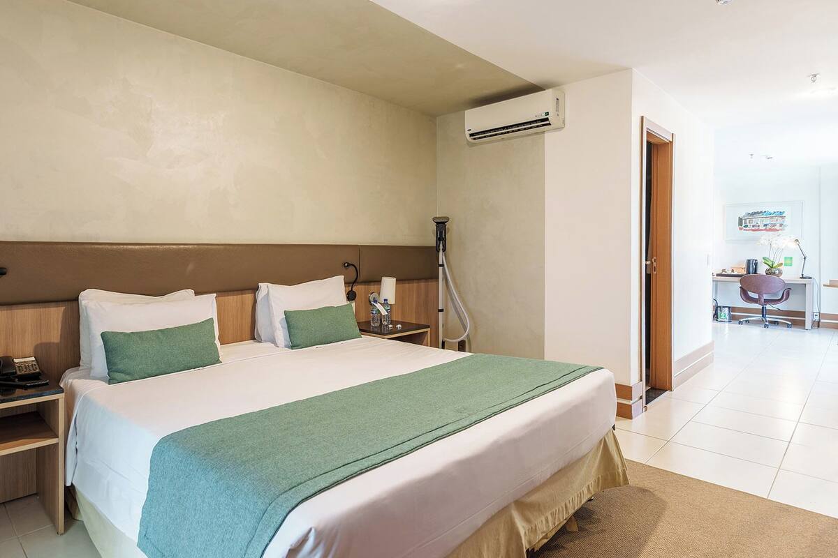 A hotel room with a bed, bedside tables, a desk, an air conditioning unit, and a bathroom visible through an open door.