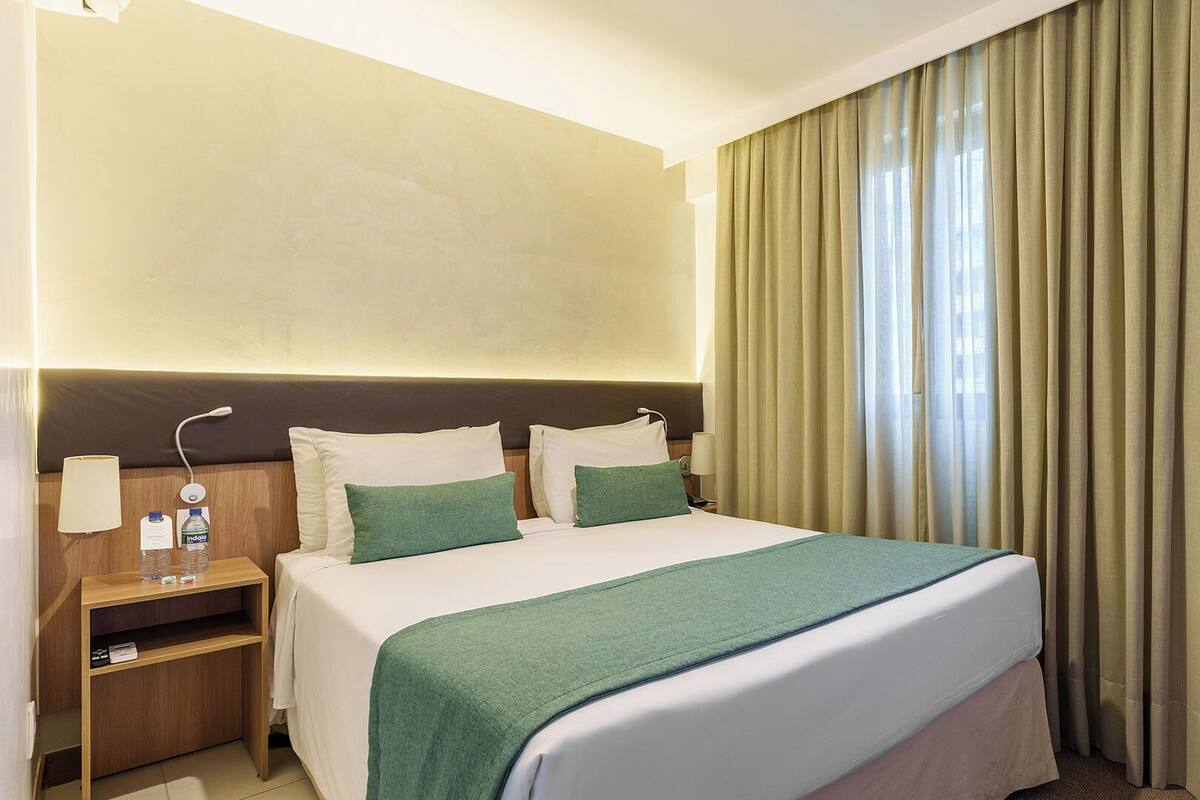 A hotel room with a bed, a nightstand, a lamp, and a window with curtains.