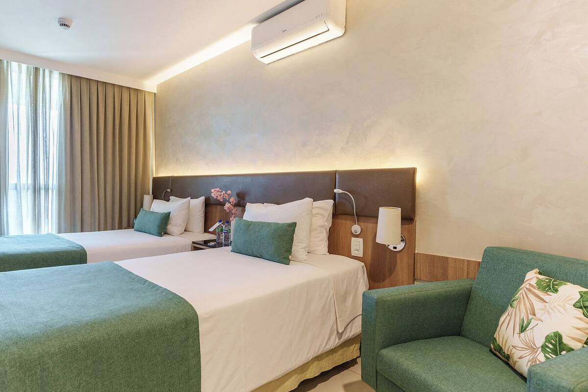 A hotel room with two beds, a sofa, a wall-mounted air conditioner, and a window with curtains.
