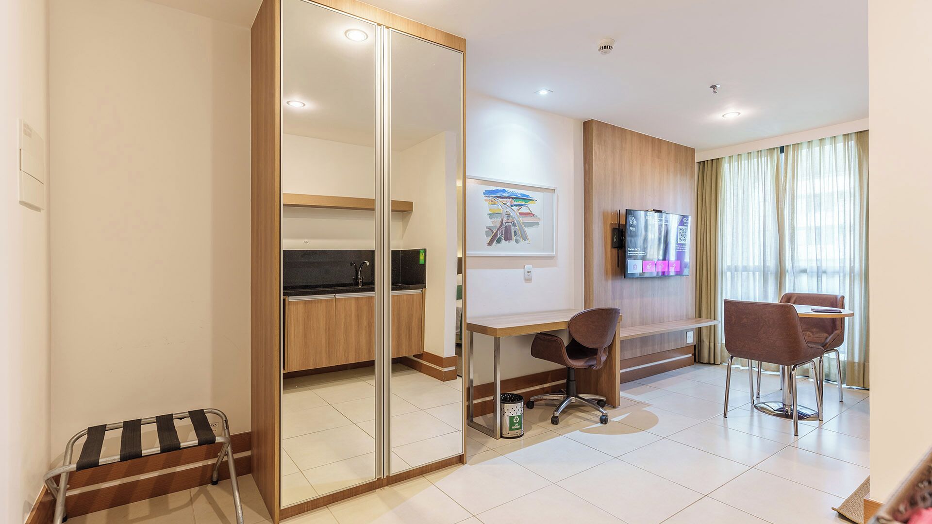 A modern hotel room with a kitchenette, a dining area with two chairs, a TV mounted on the wall, and a large window with curtains.