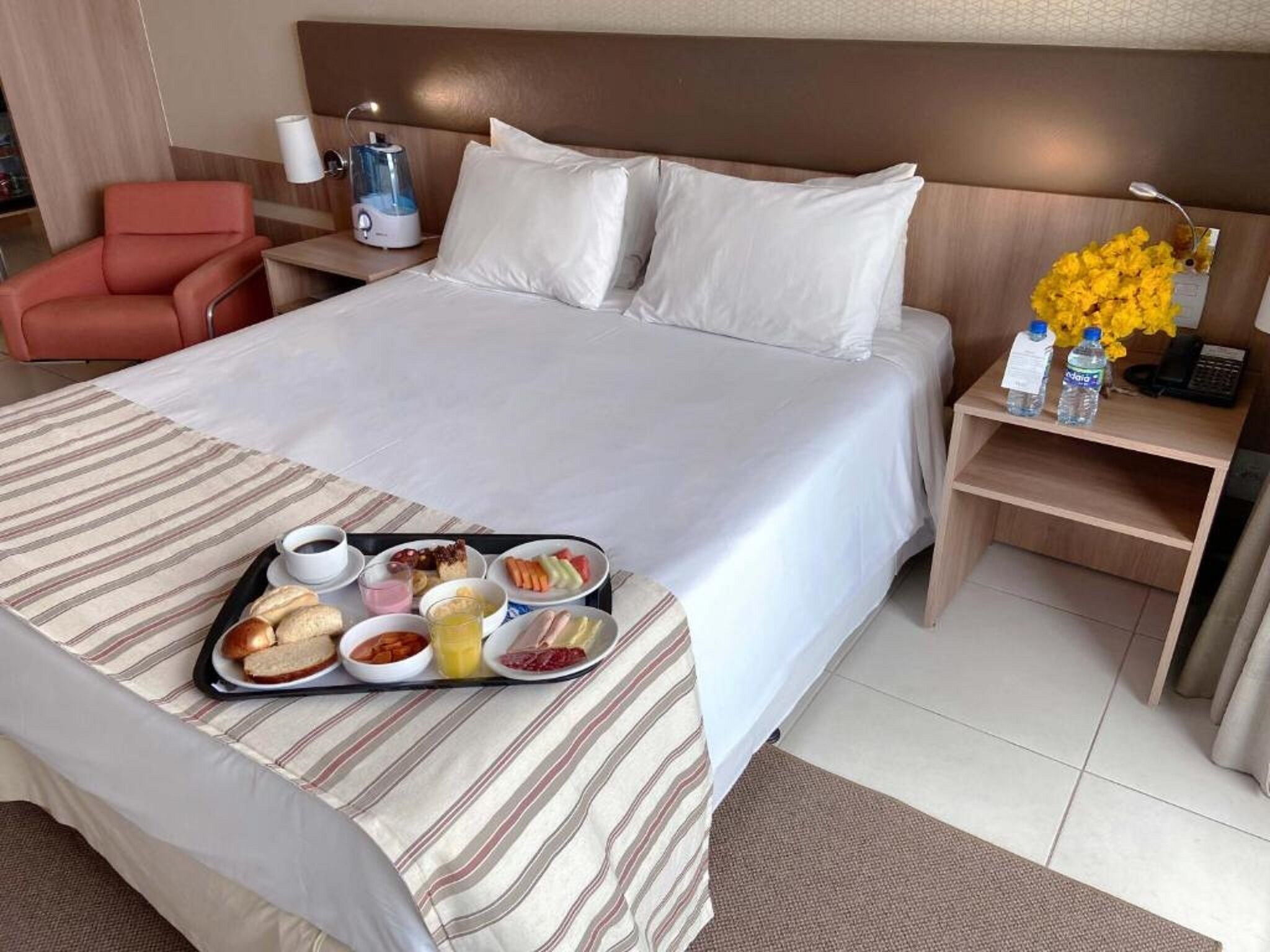 Une chambre d’hôtel avec un lit, une table de chevet avec une lampe, une chaise et un plateau contenant des articles pour le petit-déjeuner.