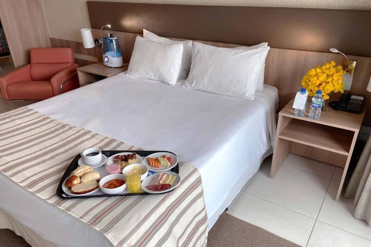 A hotel room with a bed, a nightstand with a lamp, a chair, and a tray with breakfast items.