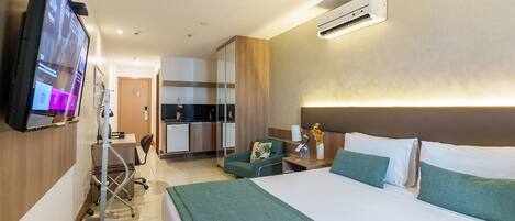 A hotel room with a bed, a desk, a chair, a TV, a kitchenette, and an air conditioning unit.