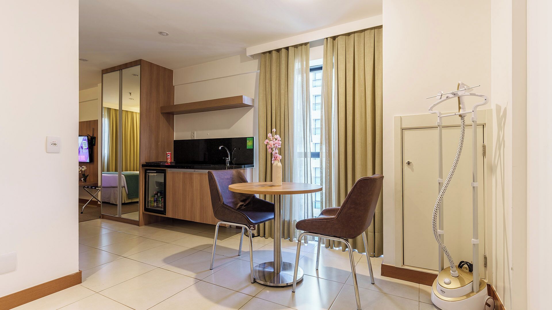 A hotel room with a kitchenette, a dining area with two chairs, a small table, a mirror, a TV, and a door with a coat rack.