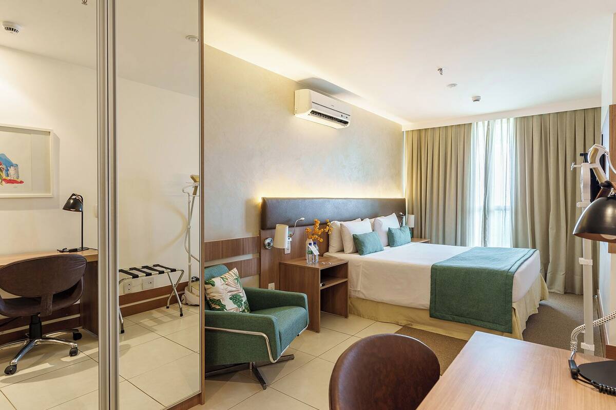 A hotel room with a bed, bedside table, lamp, chair, desk, and an air conditioning unit.