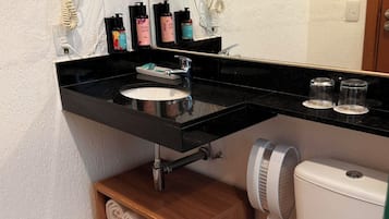 A bathroom with a black countertop, a white sink, a mirror, and a toilet. There are towels on a shelf and a trash can.