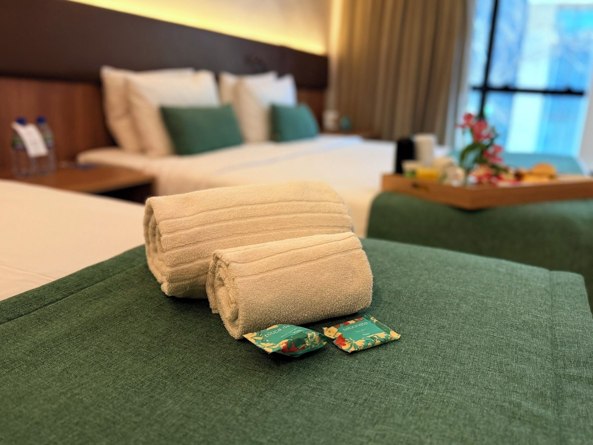 Cama bem arrumada com roupa de cama verde e duas toalhas enroladas sobre ela. Há dois pequenos sachês de higiene pessoal ao lado das toalhas.