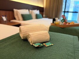 Una cama bien tendida con ropa de cama verde y dos toallas enrolladas encima. Hay dos pequeños kits de baño junto a las toallas.