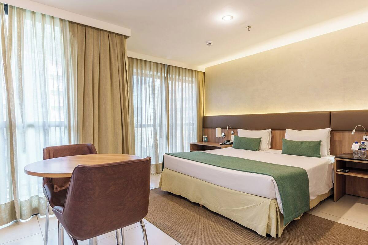 A hotel room with a bed, bedside tables, a desk, a chair, and a window with curtains.