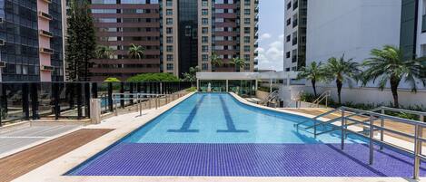 Una piscina con solárium azulejado, rodeada de edificios altos y palmeras.