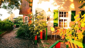 A cobblestone path leading to a rustic house with a tiled roof, a wooden deck, and a chair outside. - Hotel Restaurant Jagdhof (Steinhagen)