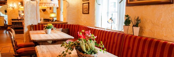 A cozy restaurant interior with wooden tables and benches, a red-striped booth, and potted plants.