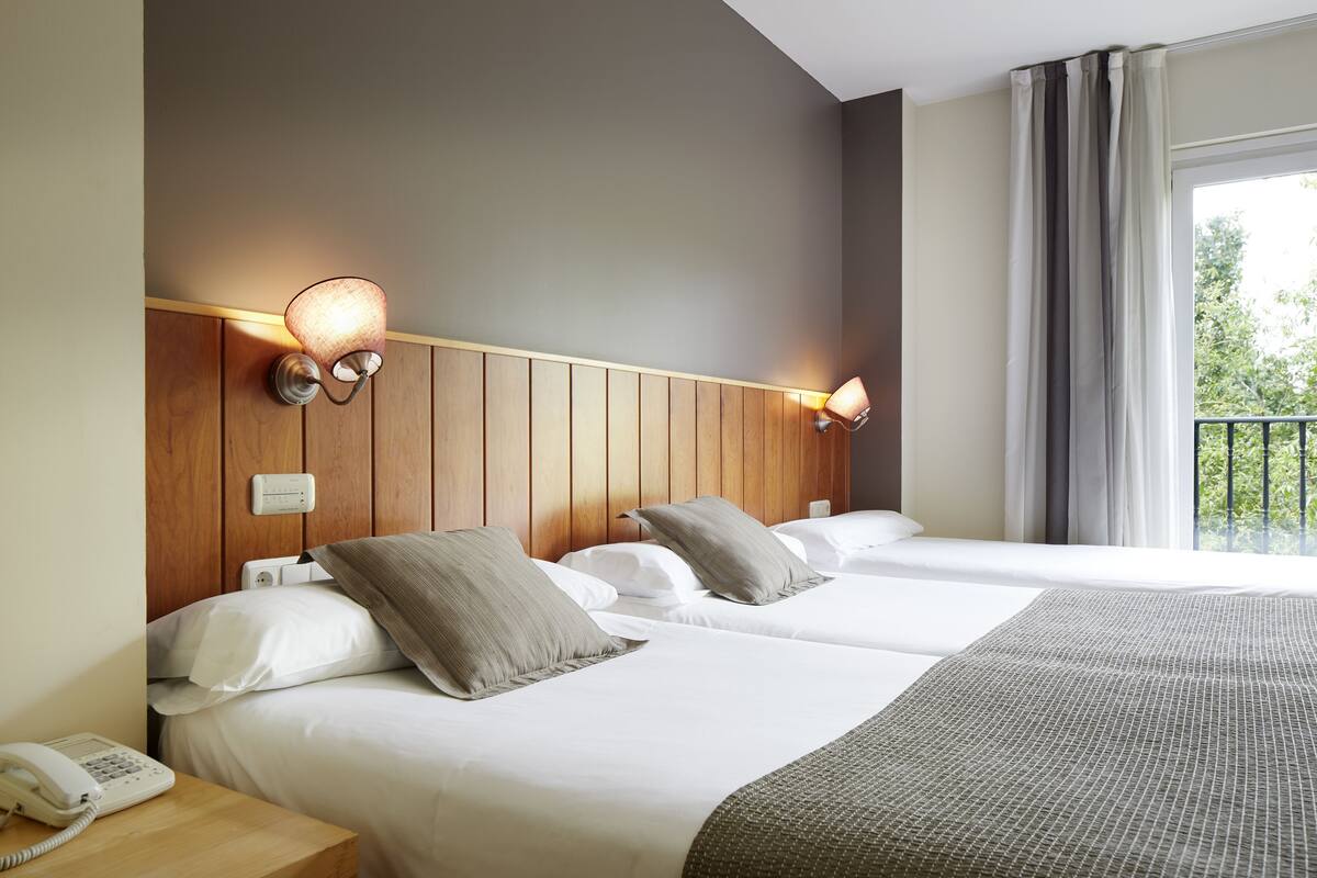 A hotel room with two beds, wooden headboard, bedside lamps, a telephone, and a window with curtains.