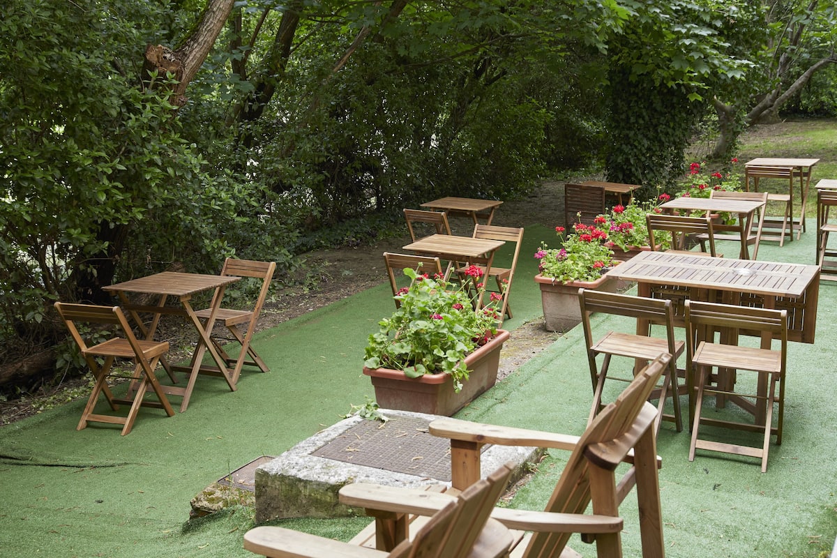 An outdoor seating area with wooden tables and chairs, surrounded by lush greenery and potted plants.