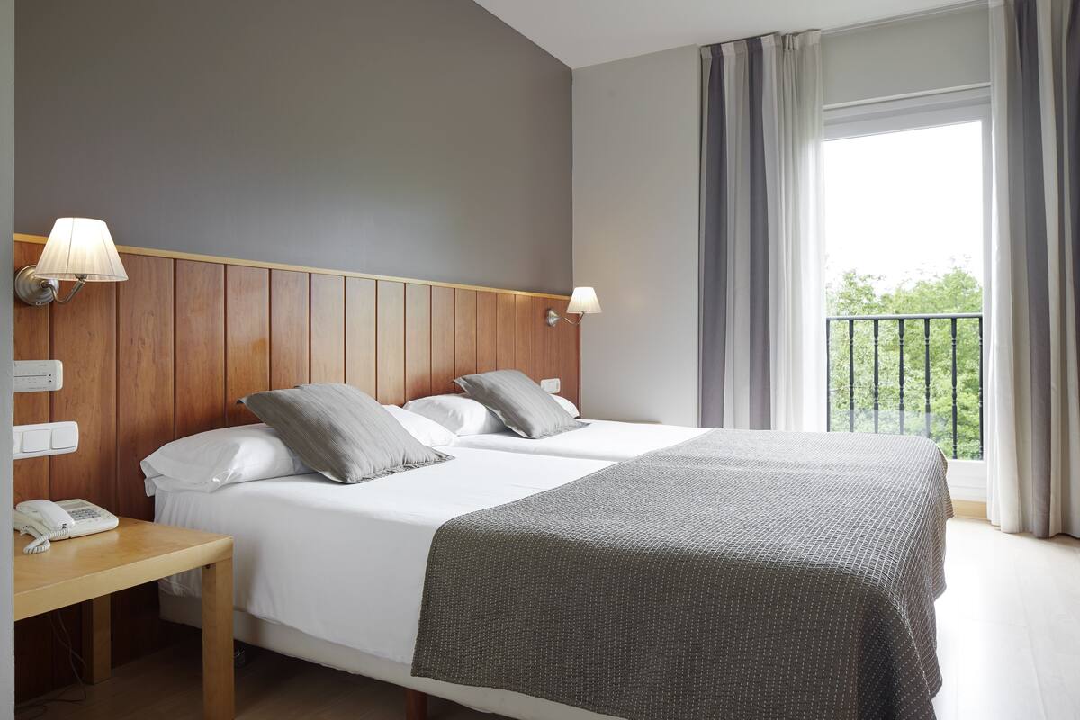 A hotel room with a double bed, two bedside lamps, a wooden headboard, a nightstand with a telephone, and a window with curtains.