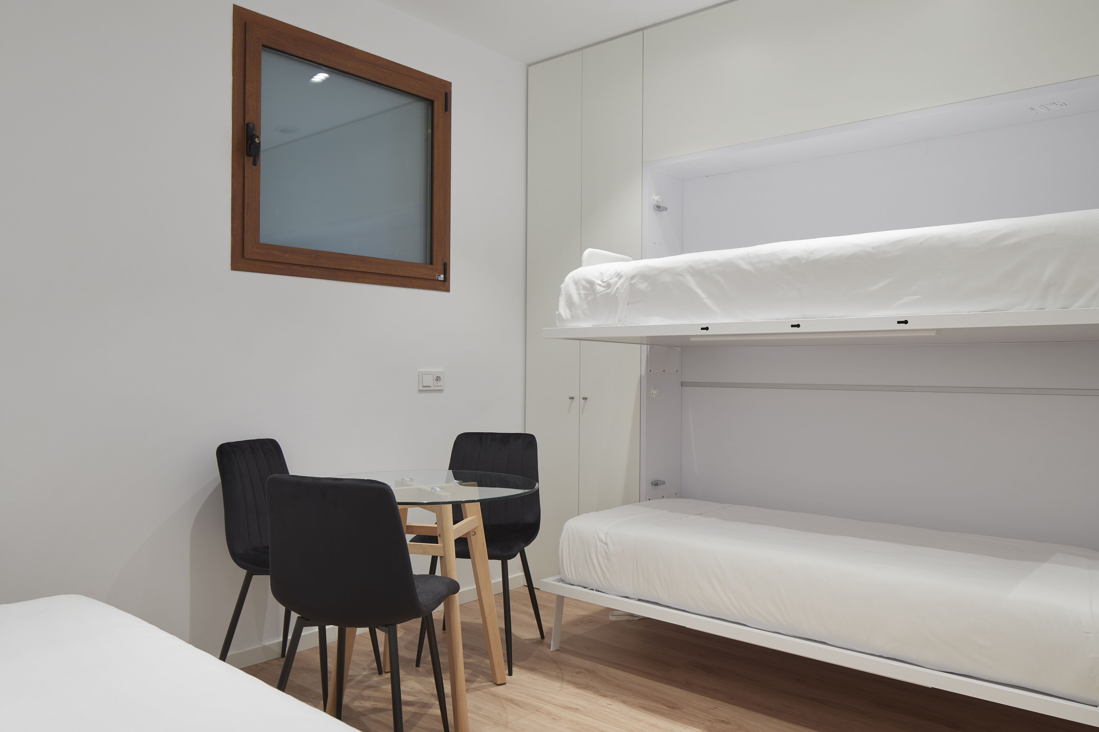 A small room with a bunk bed, a round table with two chairs, and a window with a wooden frame.
