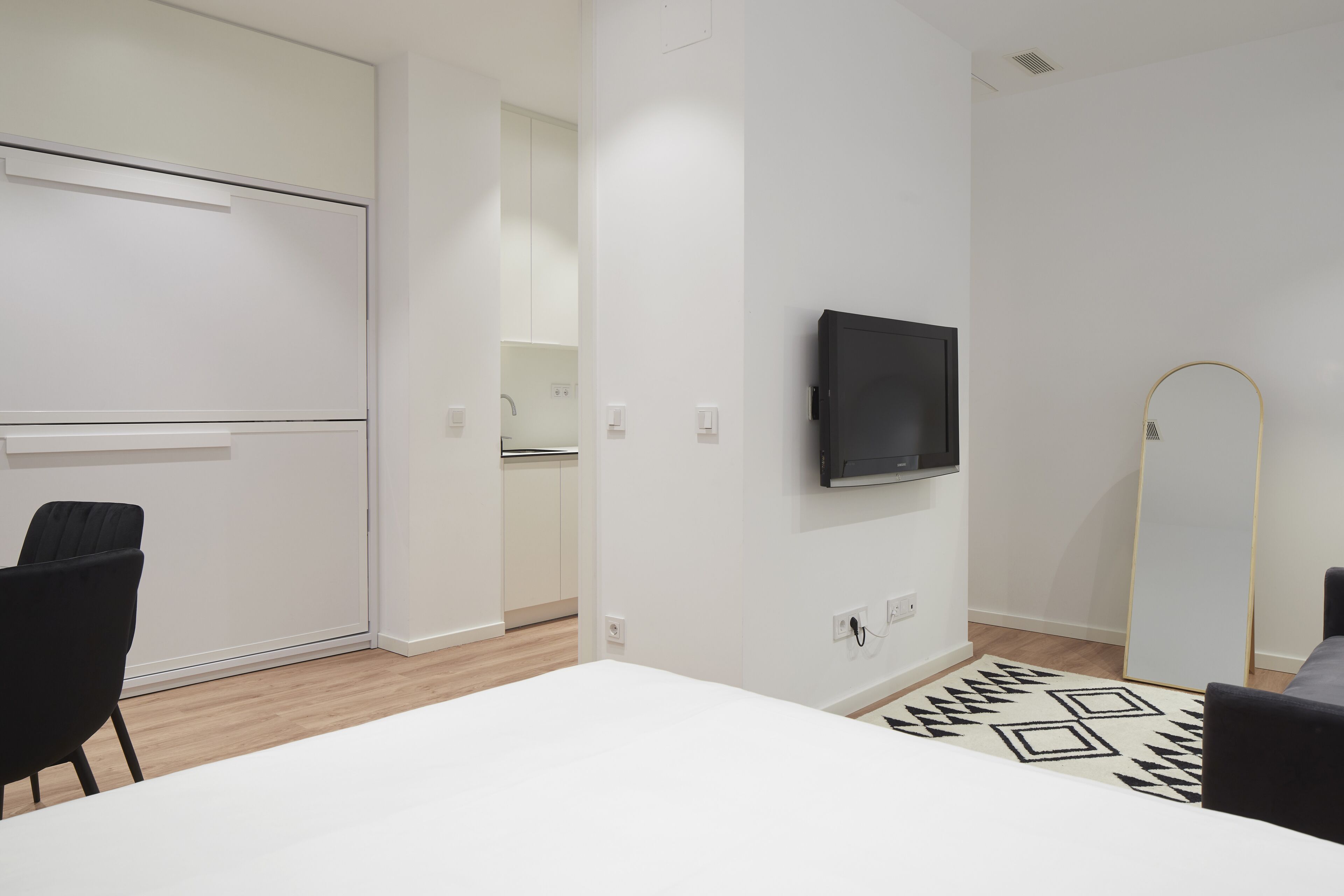 A modern, minimalist room with a white table, black chairs, a flat-screen TV, and a patterned rug.