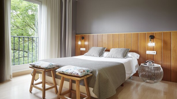 A bedroom with a large bed, two nightstands, a window with curtains, and a view of trees. - Hotel Iriguibel (Huarte)