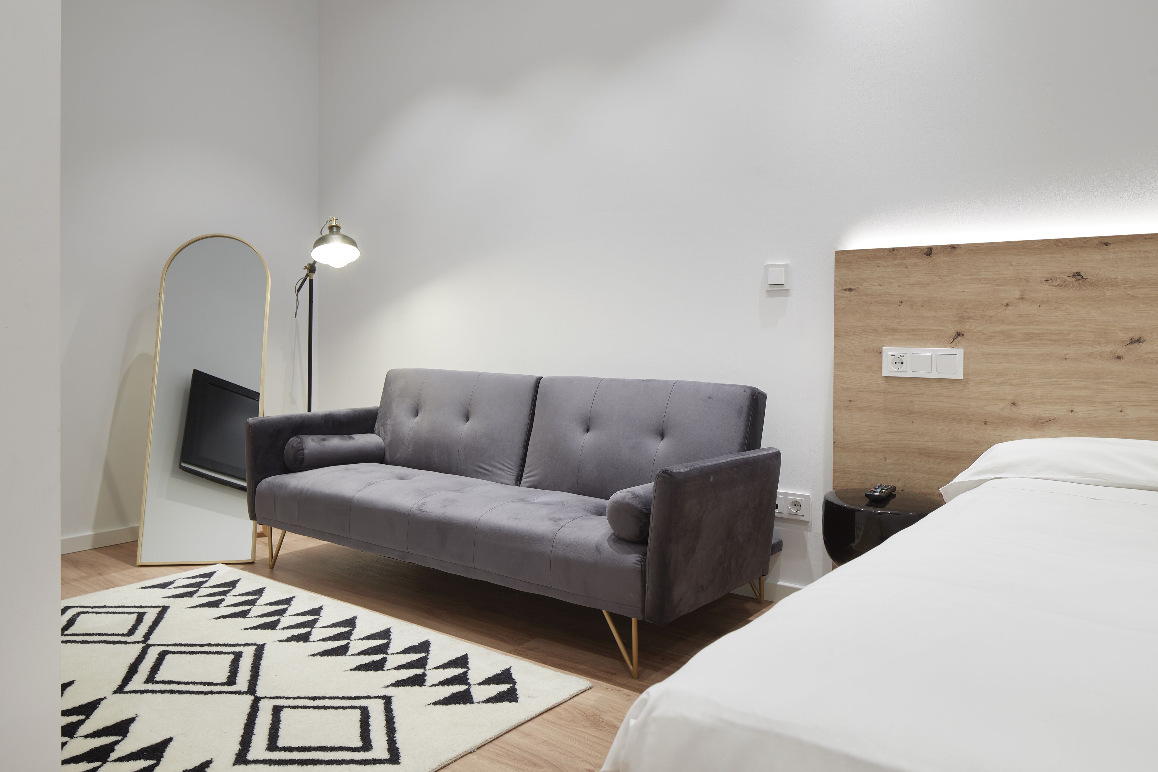 A modern bedroom with a grey sofa, a white bed, a patterned rug, a floor lamp, and a wooden headboard.