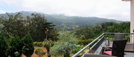 A balcony with wicker furniture, a view of a lush landscape with trees and mountains, and a clear sky.
