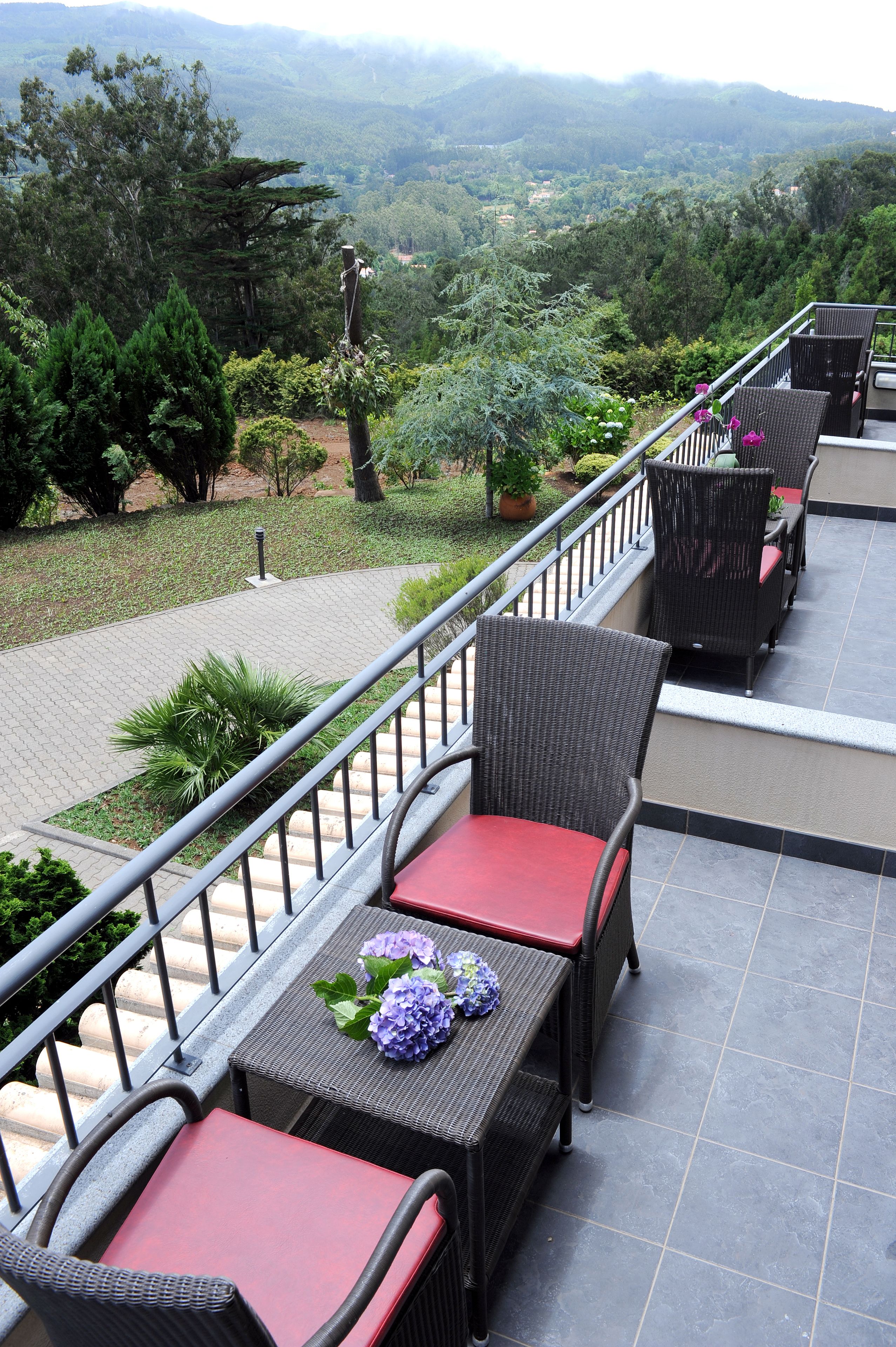 Una terrazza con mobili in vimini e cuscini rossi, con vista su un paesaggio rigoglioso, alberi e una catena montuosa in lontananza.
