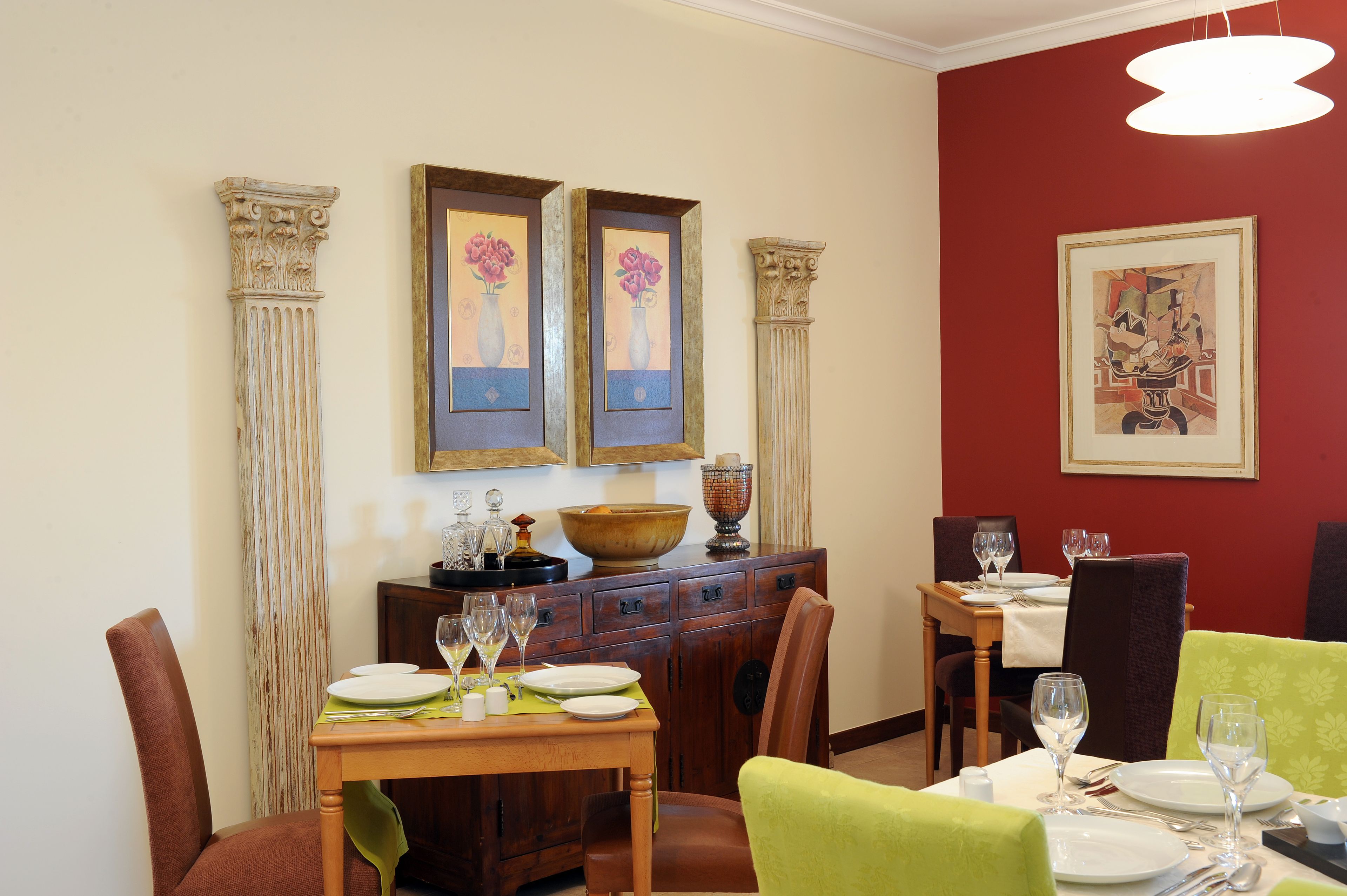 Un espace repas avec une table en bois dressée pour quatre personnes, flanquée de deux colonnes, un mur d’accent rouge et une œuvre d’art encadrée.