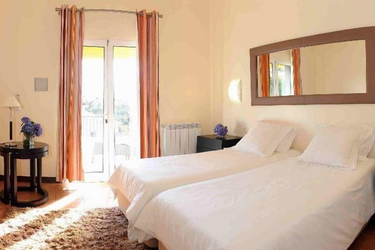A bedroom with two beds, a large mirror, a small table with a lamp, and a view of the outdoors through a door.