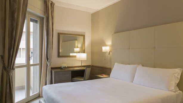 Habitación de hotel con una cama grande, un escritorio, una silla, un espejo y una ventana con cortinas.