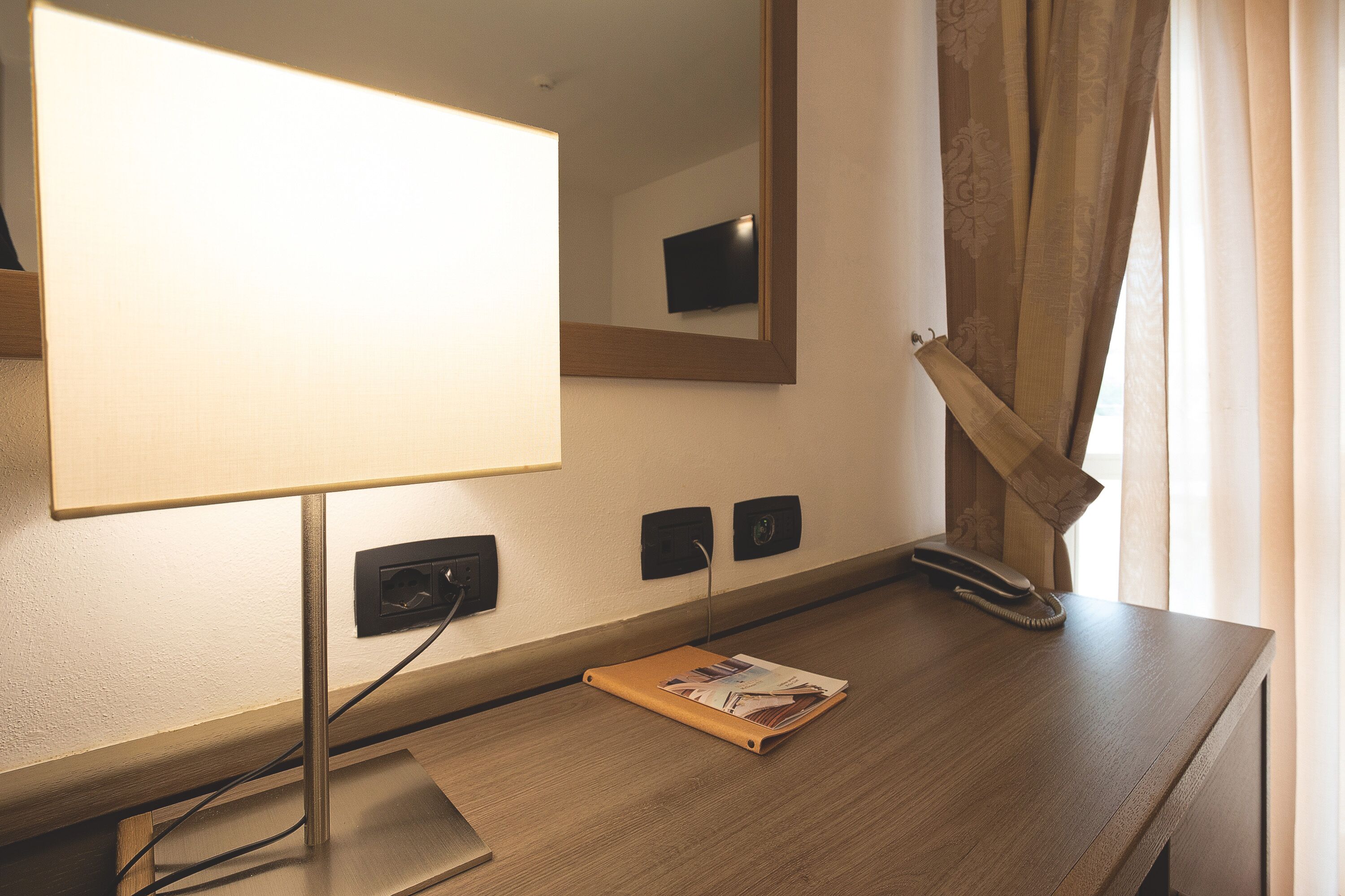 Ein Hotelzimmer mit einem Holztisch, einer Lampe, einem Spiegel, einem Telefon und einem Fenster mit Vorhängen.
