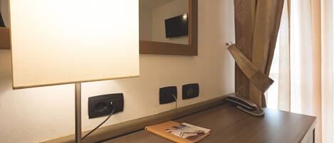 A hotel room with a wooden desk, a lamp, a mirror, a telephone, and a window with curtains.