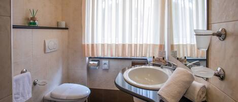 A bathroom with a toilet, bidet, sink, and mirror. There is a towel rack and a shelf with toiletries.